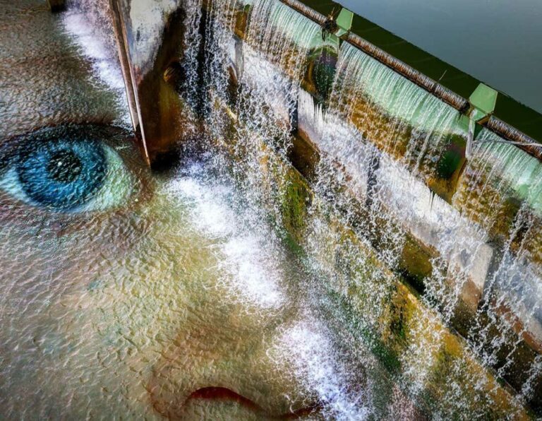 Rechts fließt Wasserfall artig Wasser von einem Damm runter und links spiegelt sich ein großes Gesicht im Wasser.