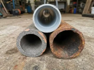 Drei Rohrquerschnitte aus PVC, Steinzeug und Gusseisen auf verwittertem Beton, Nahaufnahme mit sichtbaren Materialtexturen.